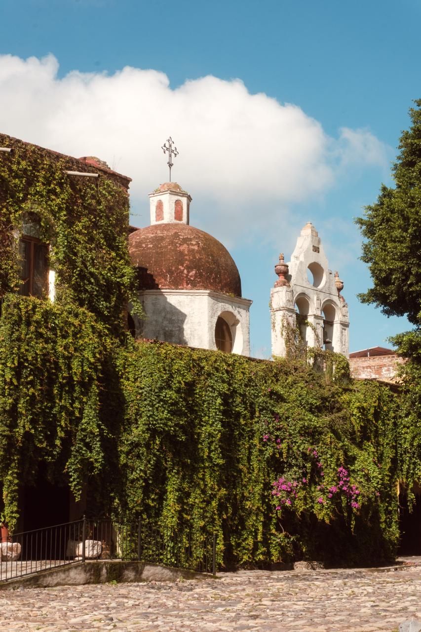 Un edificio histórico con una cúpula y torres de campanas, rodeado de muros cubiertos de hiedra y un patio empedrado.