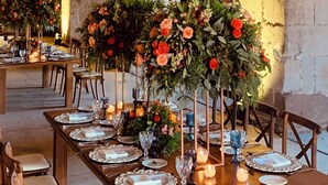 A long banquet table set with floral centerpieces, candles, and dinnerware in a rustic venue with stone arches and hanging light fixtures.
