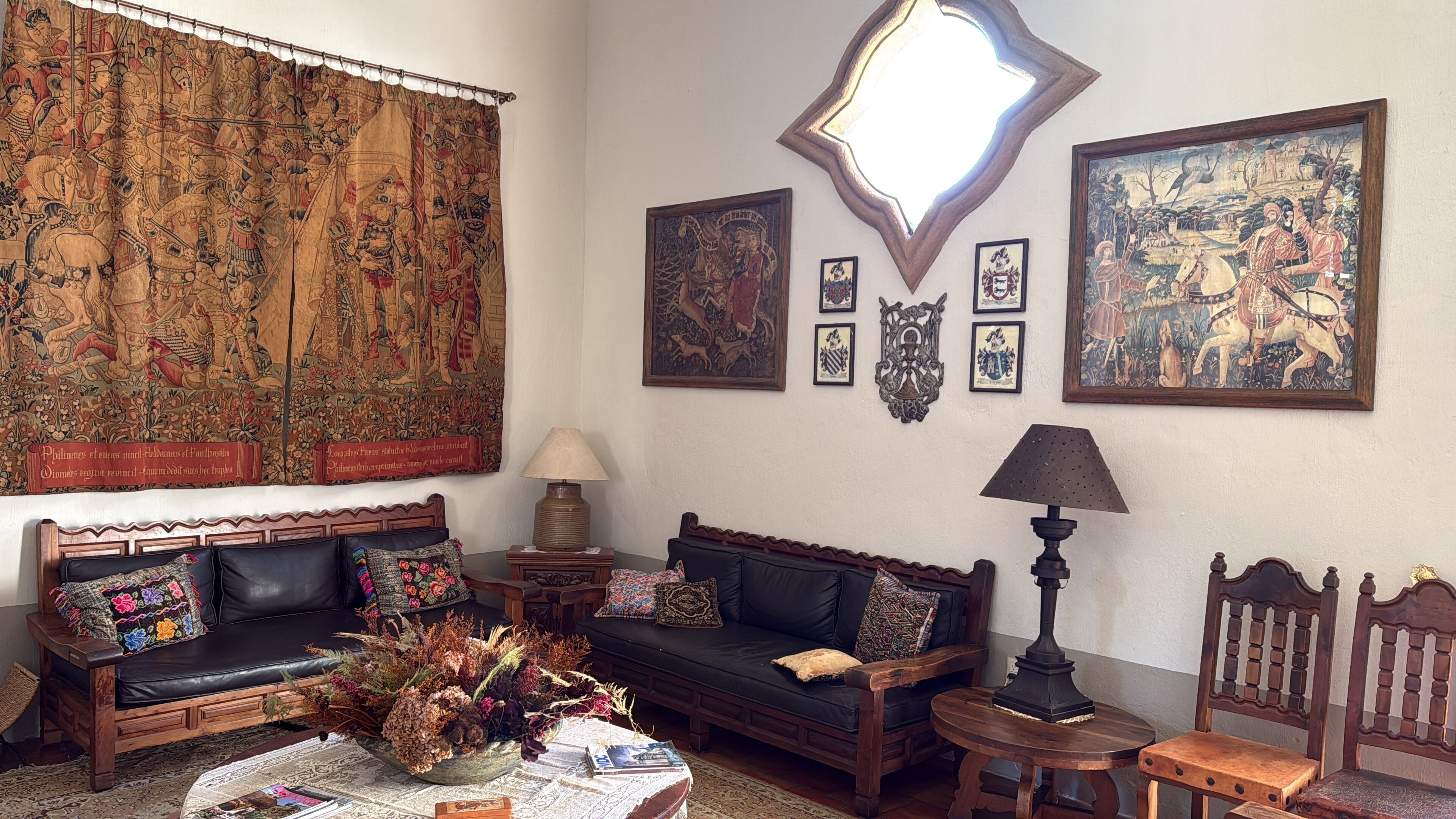 A traditional living room with a large tapestry, a sofa set, a small table with a vase of dried flowers, and a lamp.