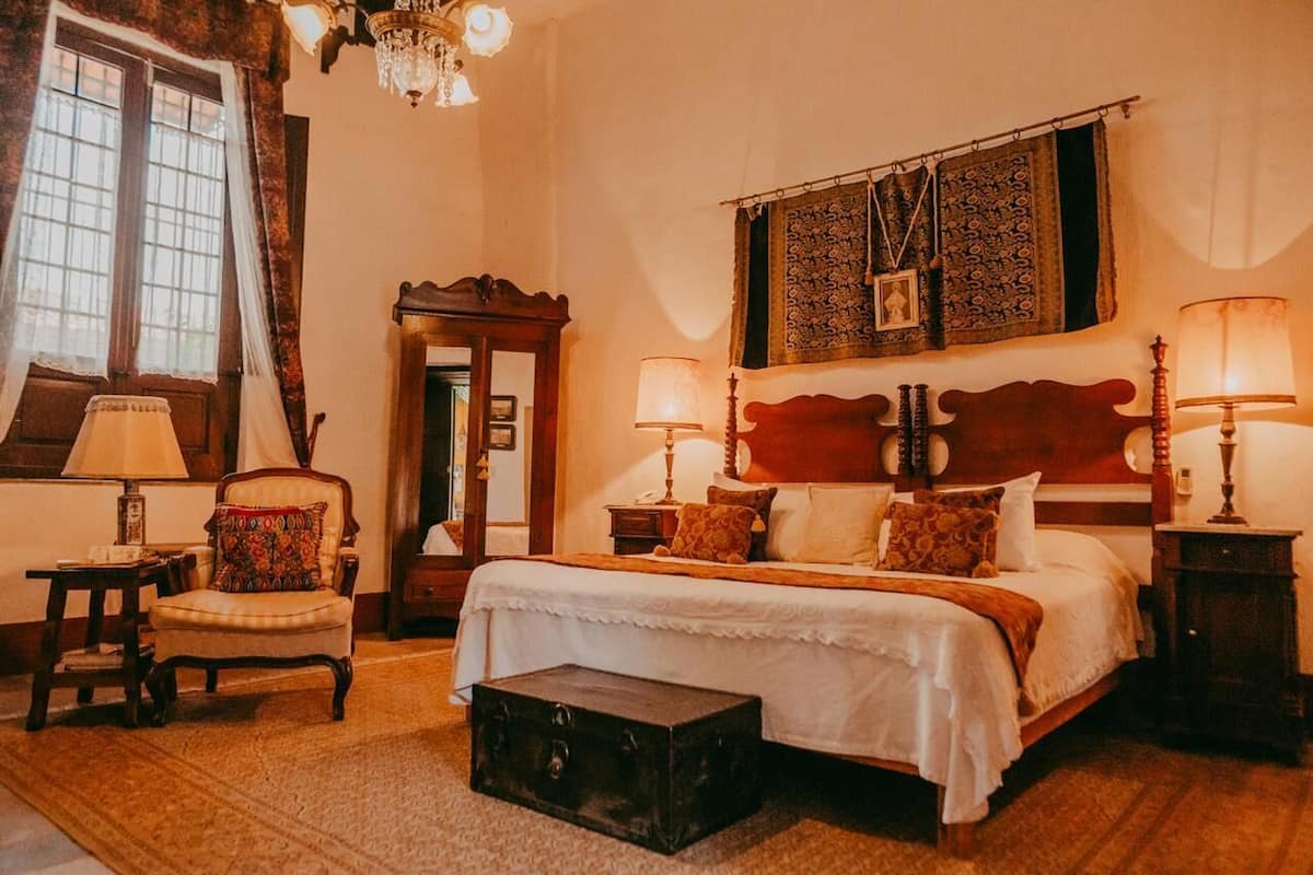 A traditional bedroom with a large bed, two bedside tables with lamps, a chair, a chest of drawers, and a mirror.