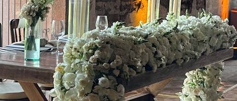 A long table with white floral arrangements and candles, set against a rustic stone wall with a warm light source.