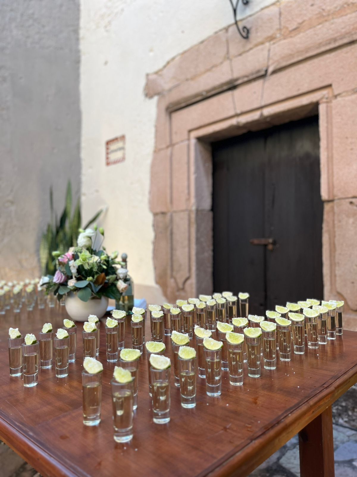 A wooden table with shot glasses filled with a clear liquid and lime slices, set against a building facade with a door and a flower arrangement.
