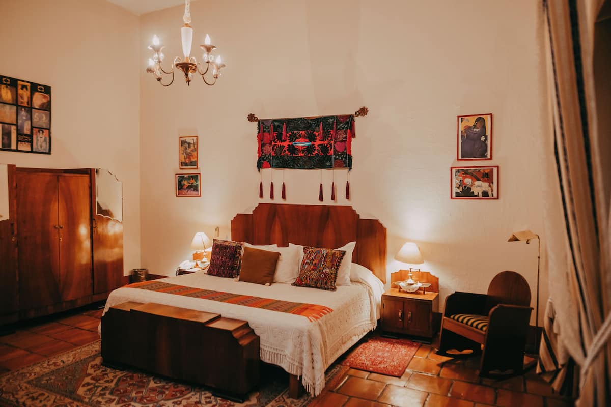A bedroom with a wooden bed, a nightstand, a chair, a chandelier, and artwork on the walls.