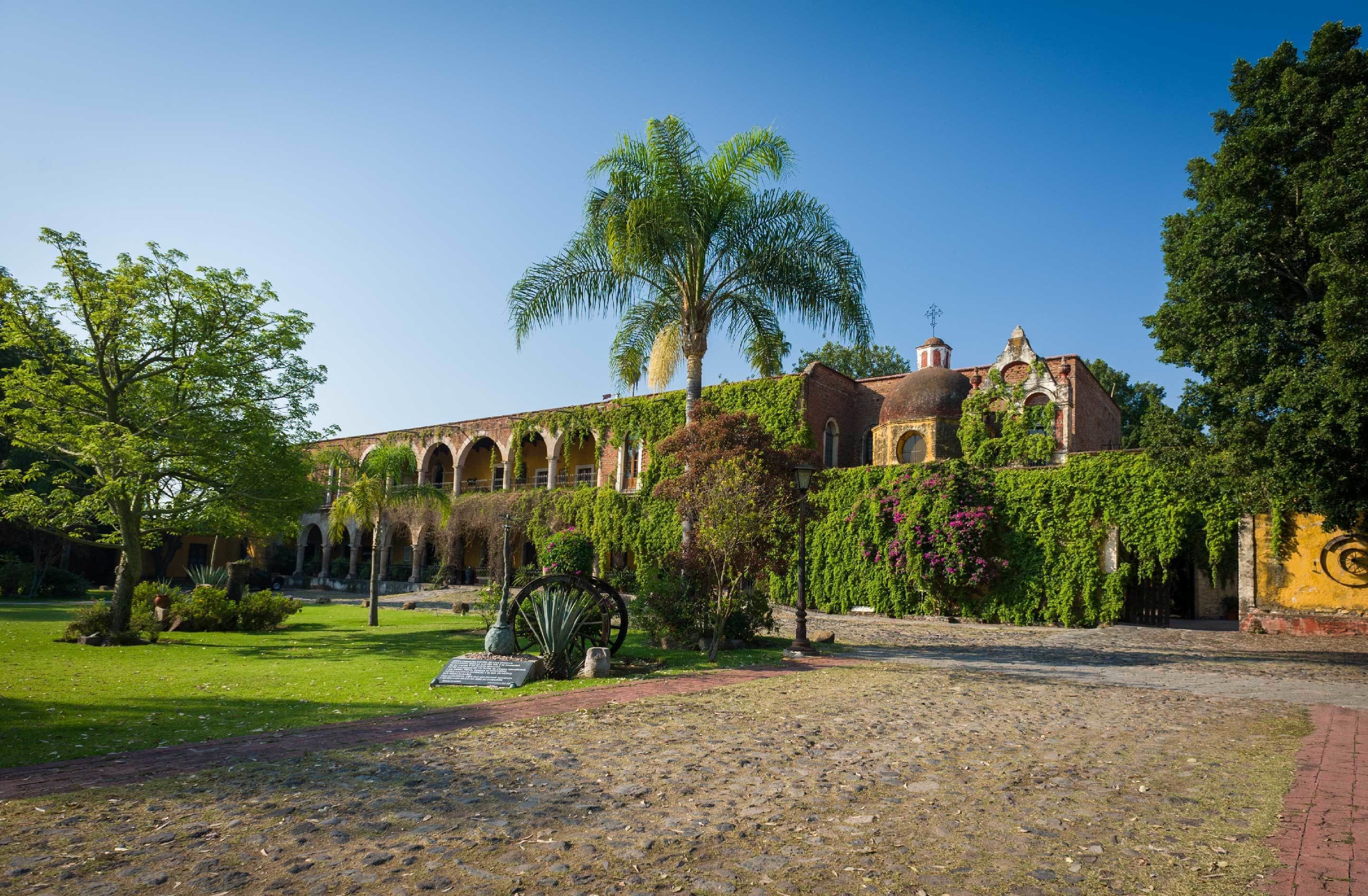 A historic building with arched windows and a central dome, surrounded by lush greenery and a cobblestone path.