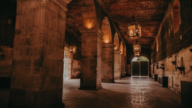 A dimly lit corridor with stone columns and hanging lanterns, leading to a large arched doorway.