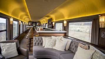 Interior de un tren con un sofá curvo, zona de bar y ventanas amplias que dan a vistas del desierto.