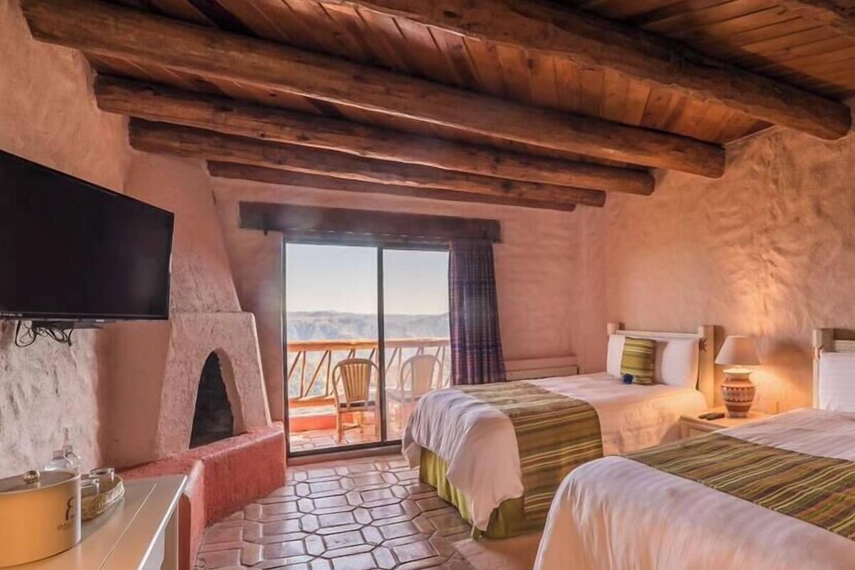 A rustic room with a fireplace, two beds, a TV, and a balcony with a view.