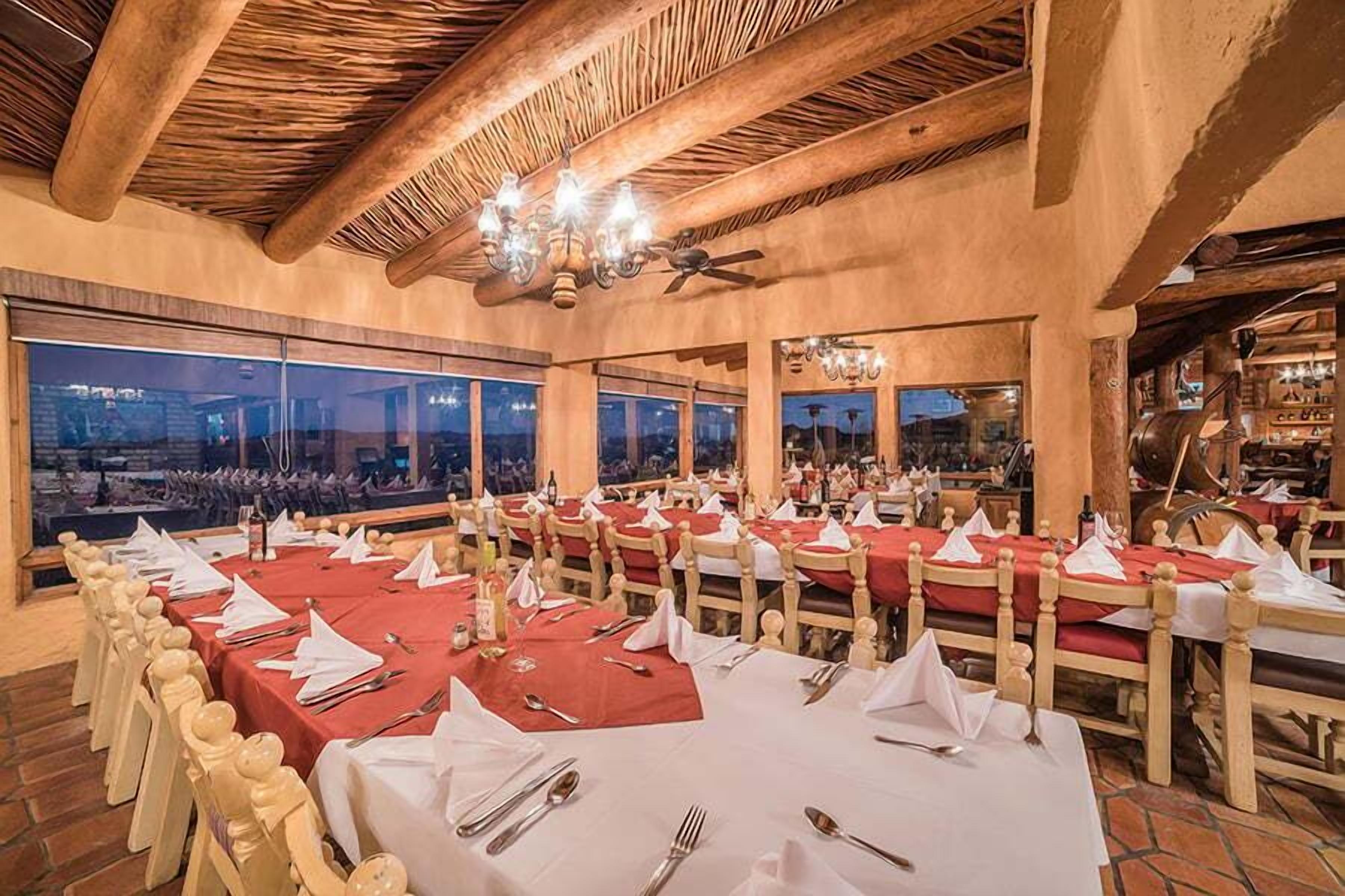 A restaurant with multiple tables set for dining, large windows offering a view, and a wooden ceiling.