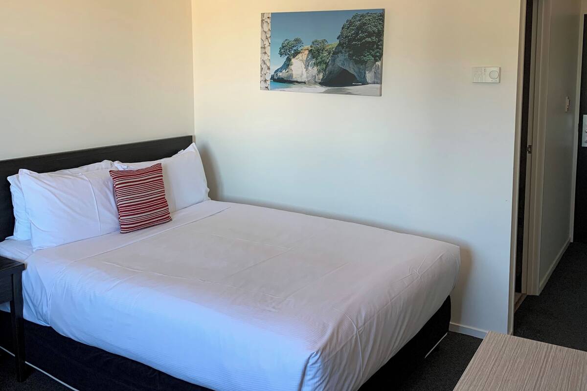 A neatly made bed with white bedding and a single red and white striped pillow. A painting of a coastal scene hangs above the bed.