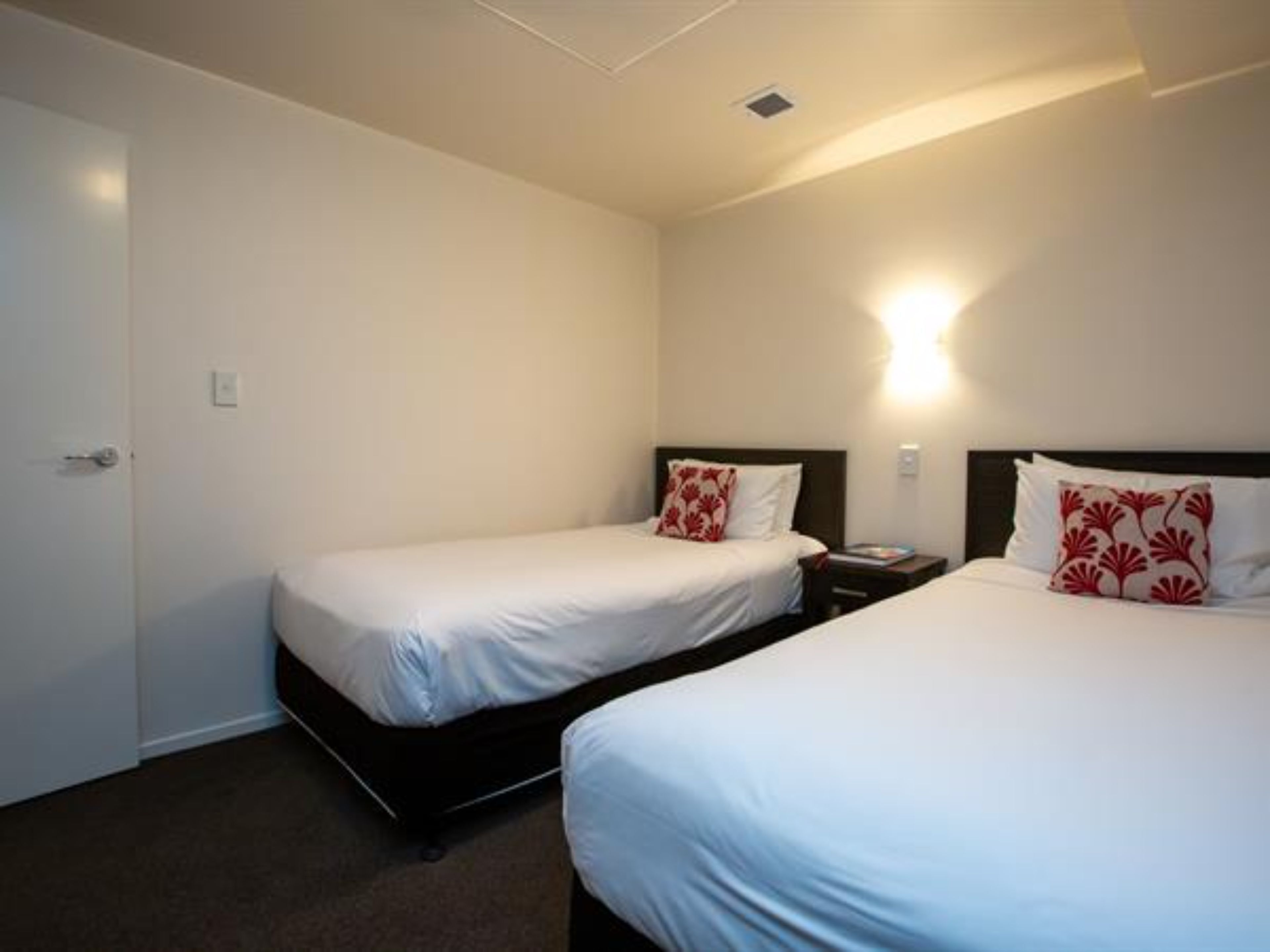 Habitación de hotel con dos camas individuales, una mesita de noche, una lámpara en la pared y una puerta.