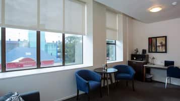 A modern living room with a sofa, two blue armchairs, a small round table, and a desk with a computer. Large windows provide a view of the outside.