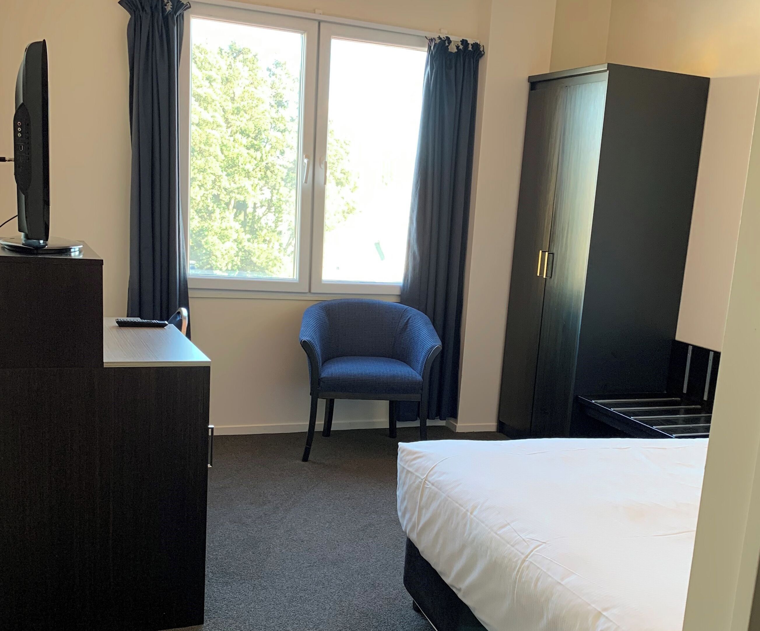 A hotel room with a bed, a chair, a desk with a television, a wardrobe, and a window with curtains.