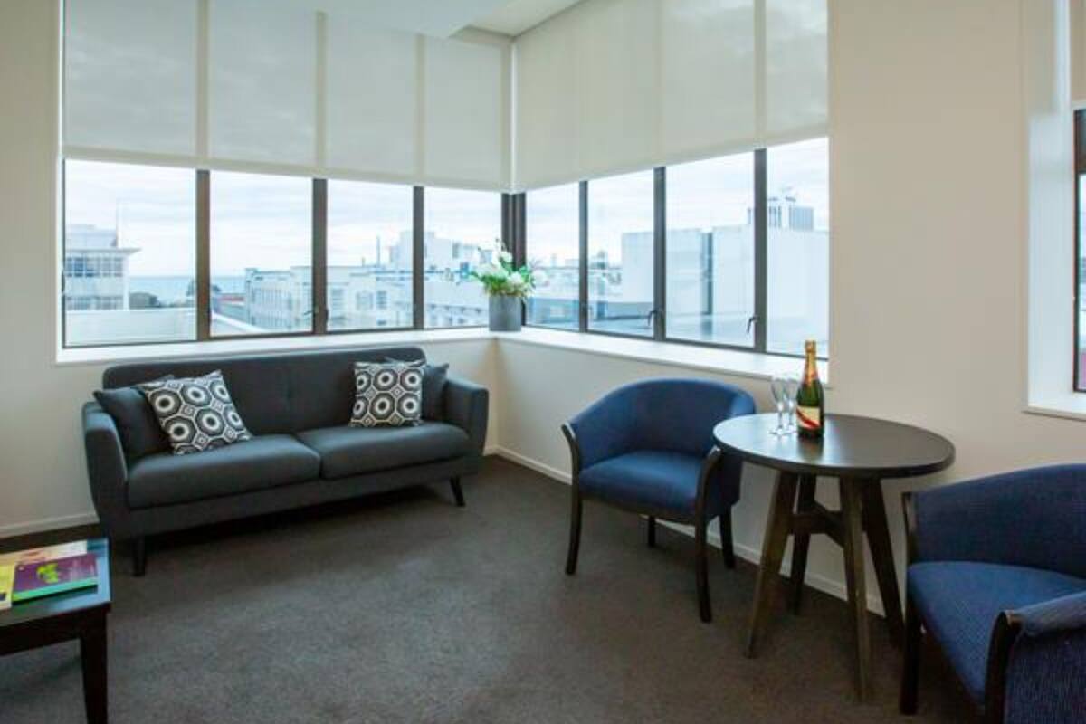 Ein modernes Zimmer mit grauem Sofa, blauem Sessel und einem kleinen runden Tisch mit einer Flasche. Große Fenster bieten Blick auf die Stadt.
