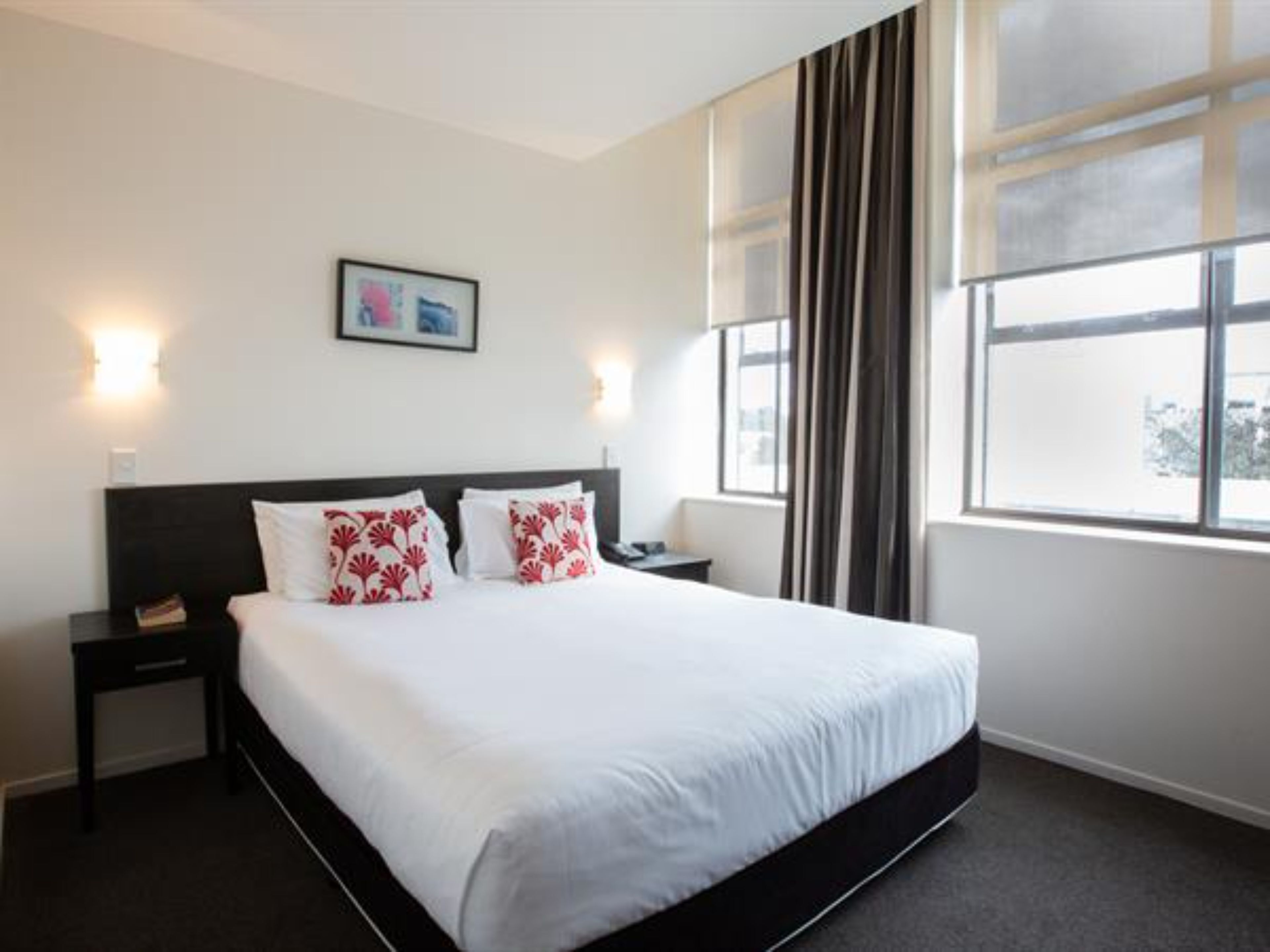 A hotel room with a large bed, a nightstand, a bedside table, a framed picture on the wall, and a window with curtains.