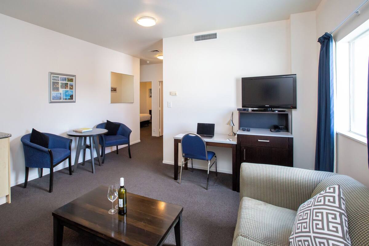 Ein kompaktes Hotelzimmer mit Küchenzeile, einer Essecke mit zwei Stühlen und einem Tisch, einem separaten Schreibtisch mit Stuhl, einem Fernseher und einem Fenster mit Vorhängen.