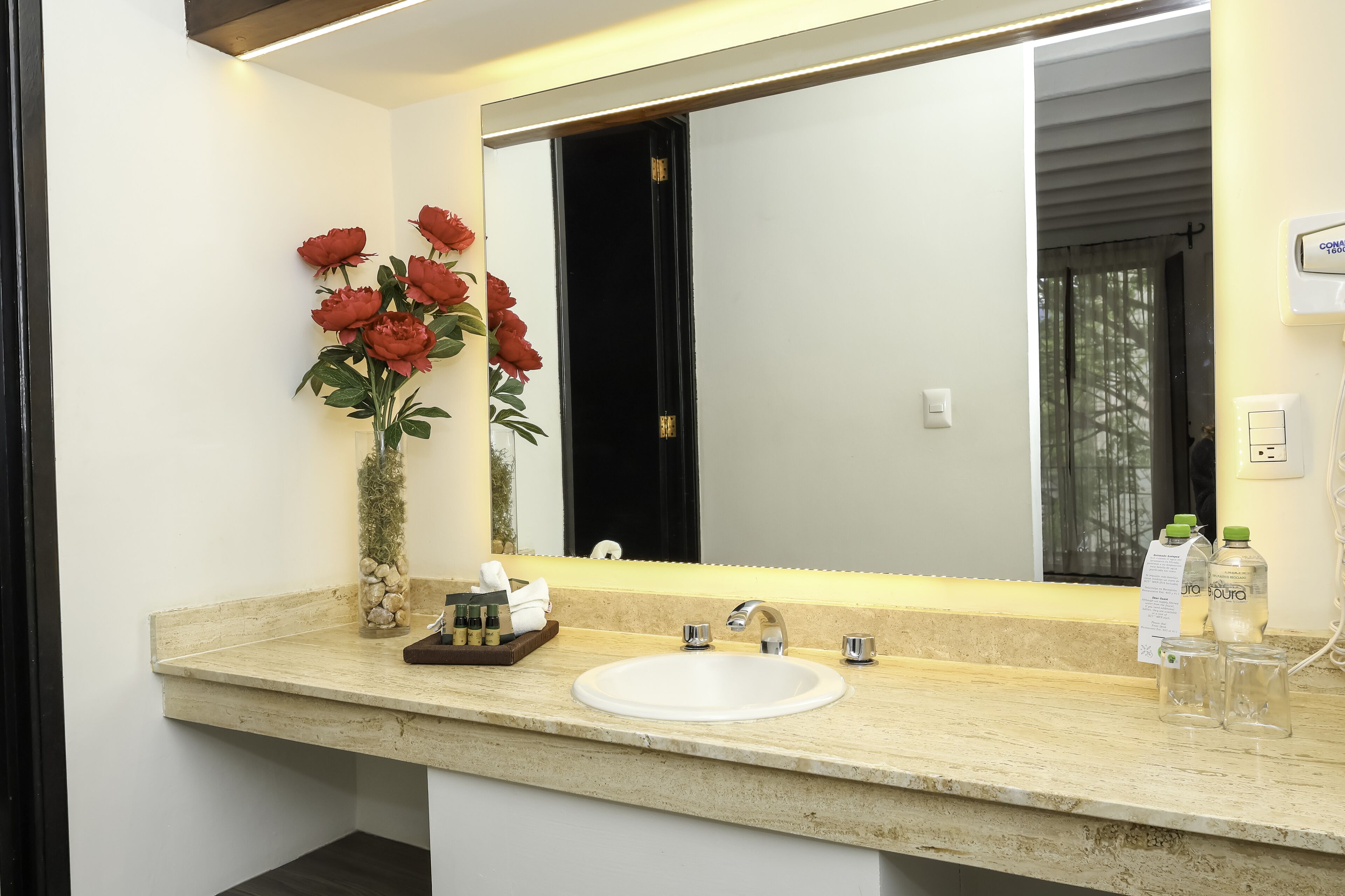 A bathroom with a white sink, a large mirror, and a vase with red flowers.