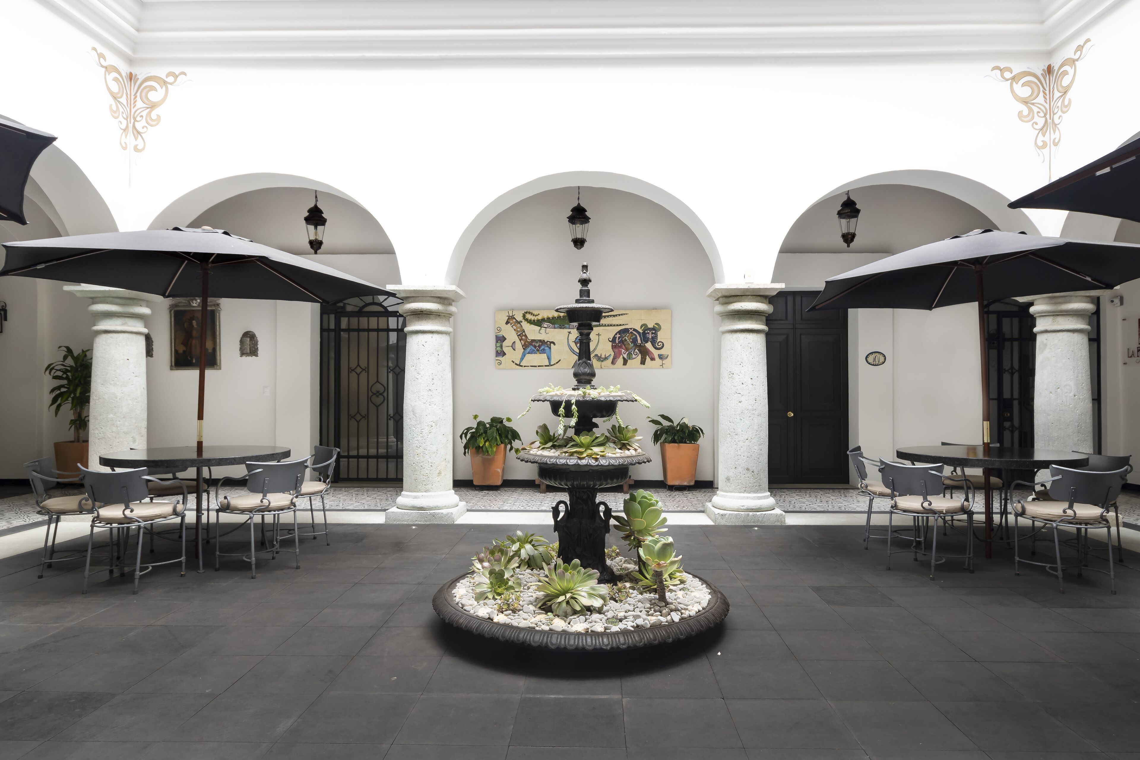 A courtyard with a central fountain, surrounded by tables and chairs, under arched ceilings with decorative wall art.
