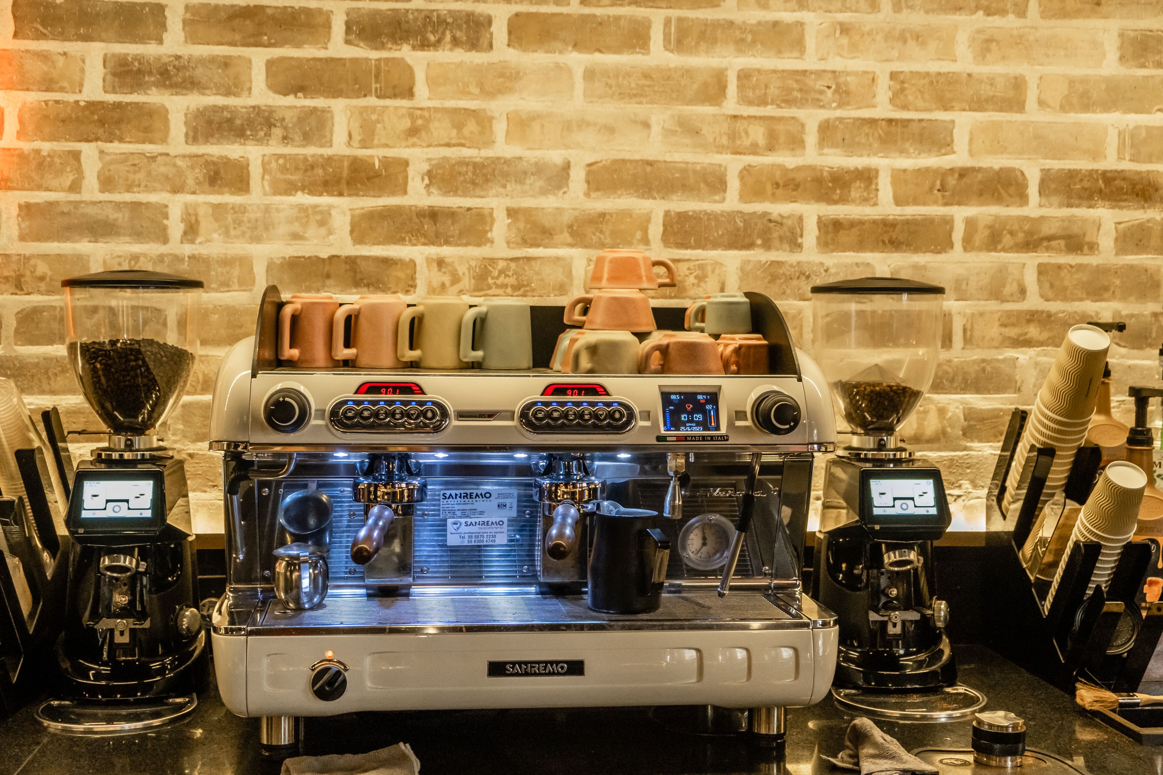 A coffee machine with multiple portafilters and a digital display, set against a brick wall.