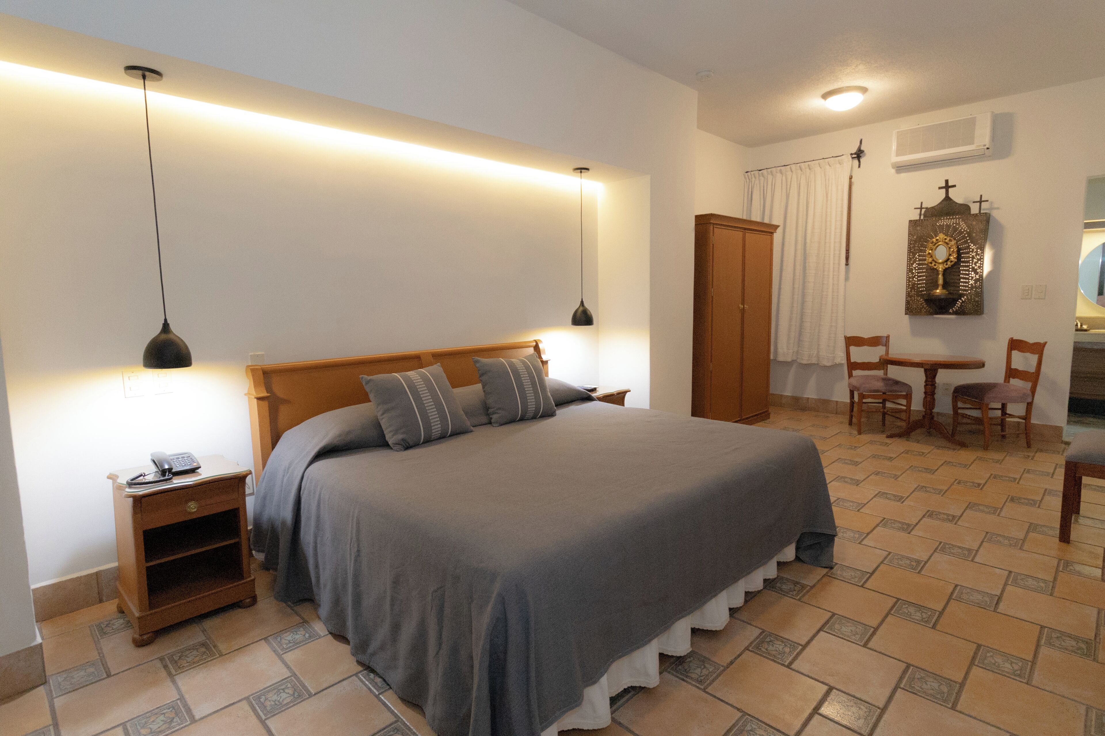 Un dormitorio ordenado con una cama grande, mesitas de noche, un armario de madera, una pequeña mesa con sillas y un equipo de aire acondicionado de pared.