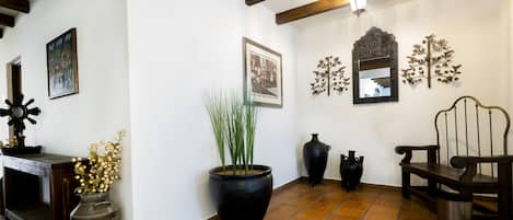 Una habitación con suelo de baldosas, vigas de madera, una olla negra grande con plantas, un banco de madera y adornos colgados en la pared.