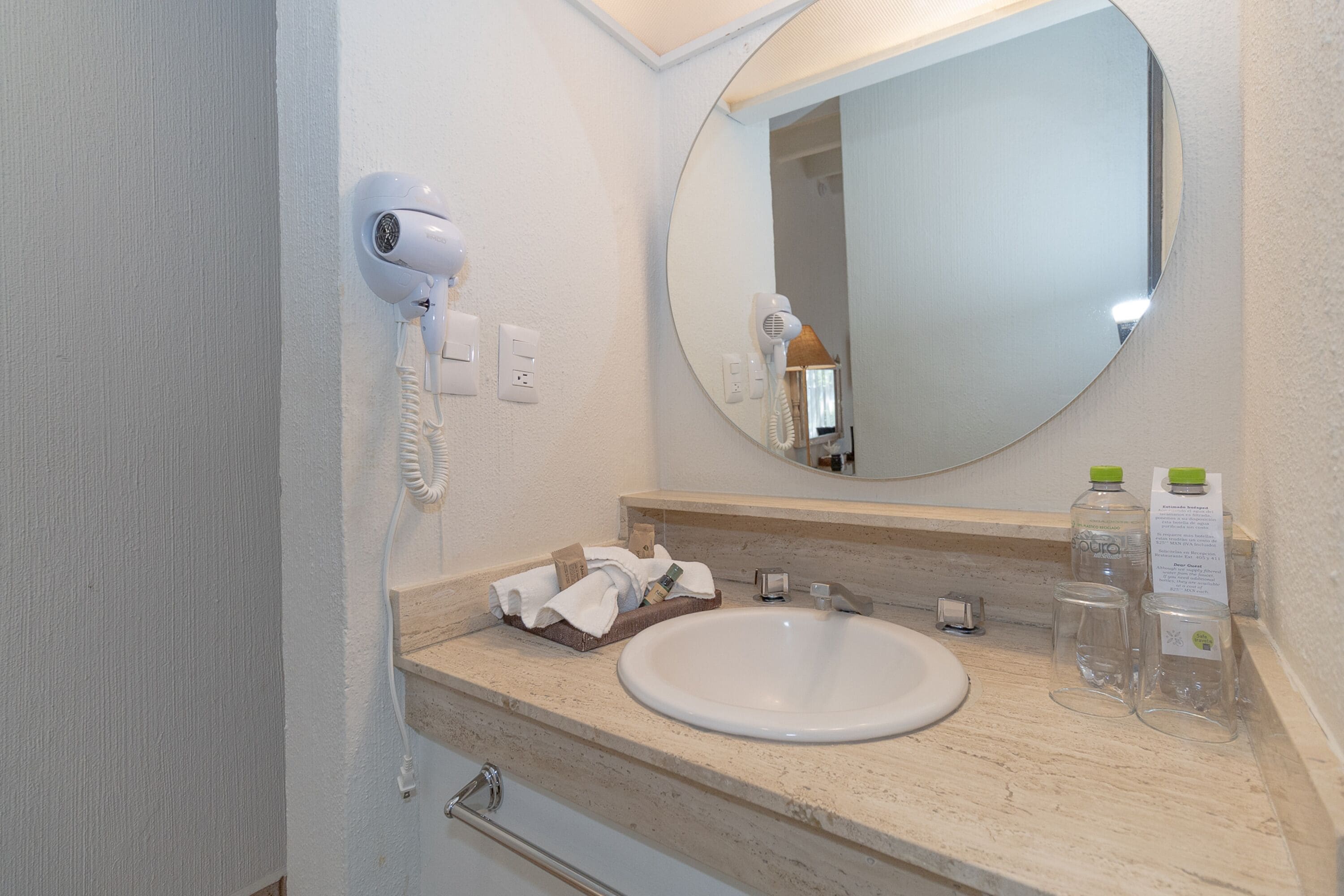 A bathroom with a white sink, a round mirror, a hairdryer, and toiletries on the counter.