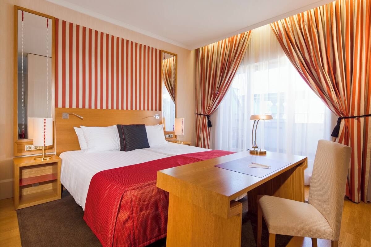 A hotel room with a large bed, a wooden desk, a chair, a nightstand with a lamp, and a mirror.