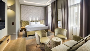 A hotel room with a large bed, two armchairs, a sofa, a wooden desk, and a large window with curtains.