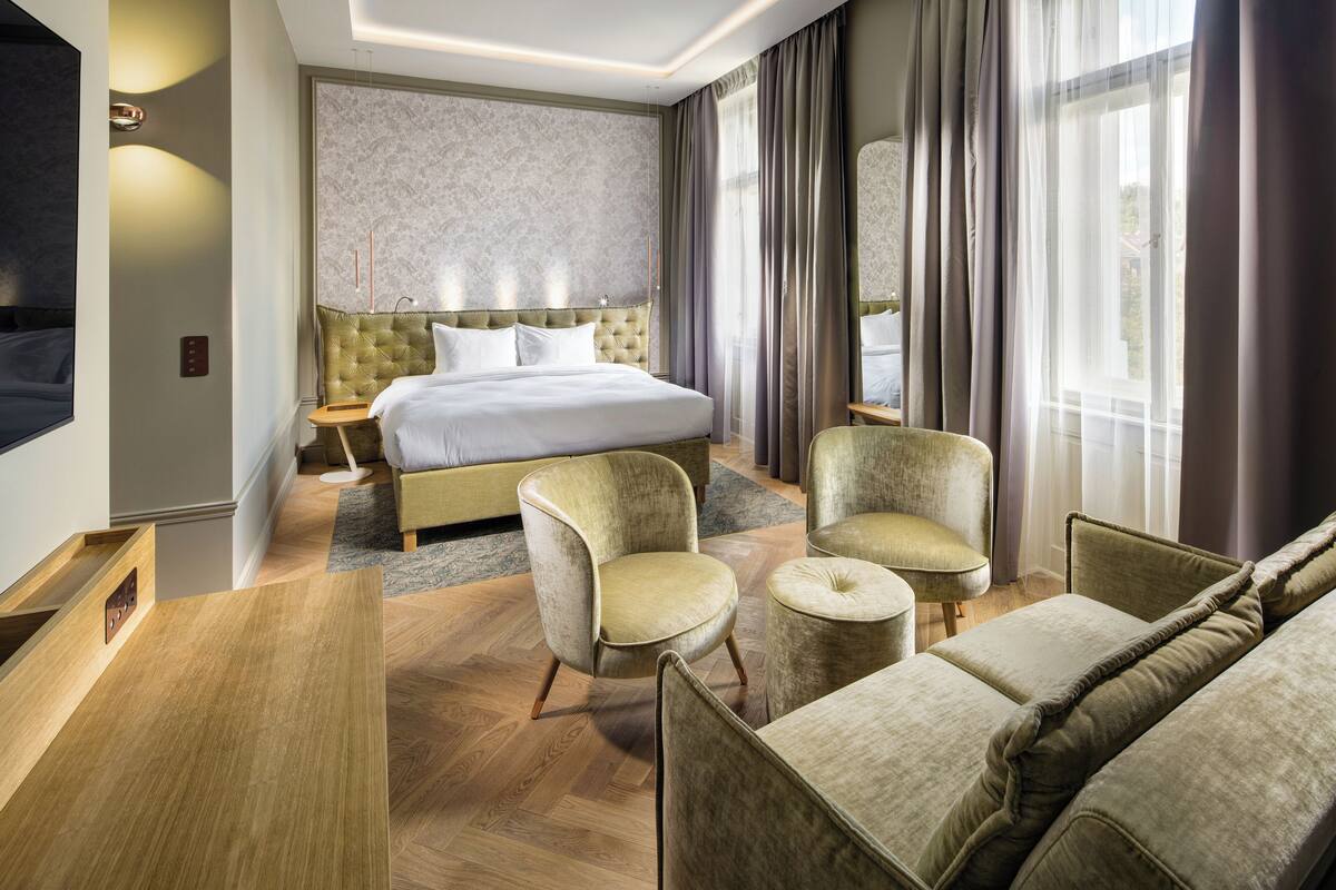 A hotel room with a large bed, two armchairs, a sofa, a wooden desk, and a large window with curtains.