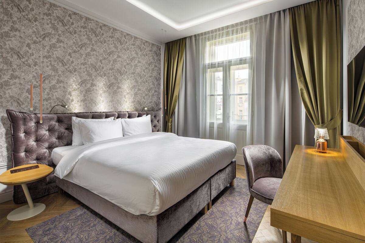 A modern hotel room with a tufted headboard bed, a wooden side table, a desk, and a chair.