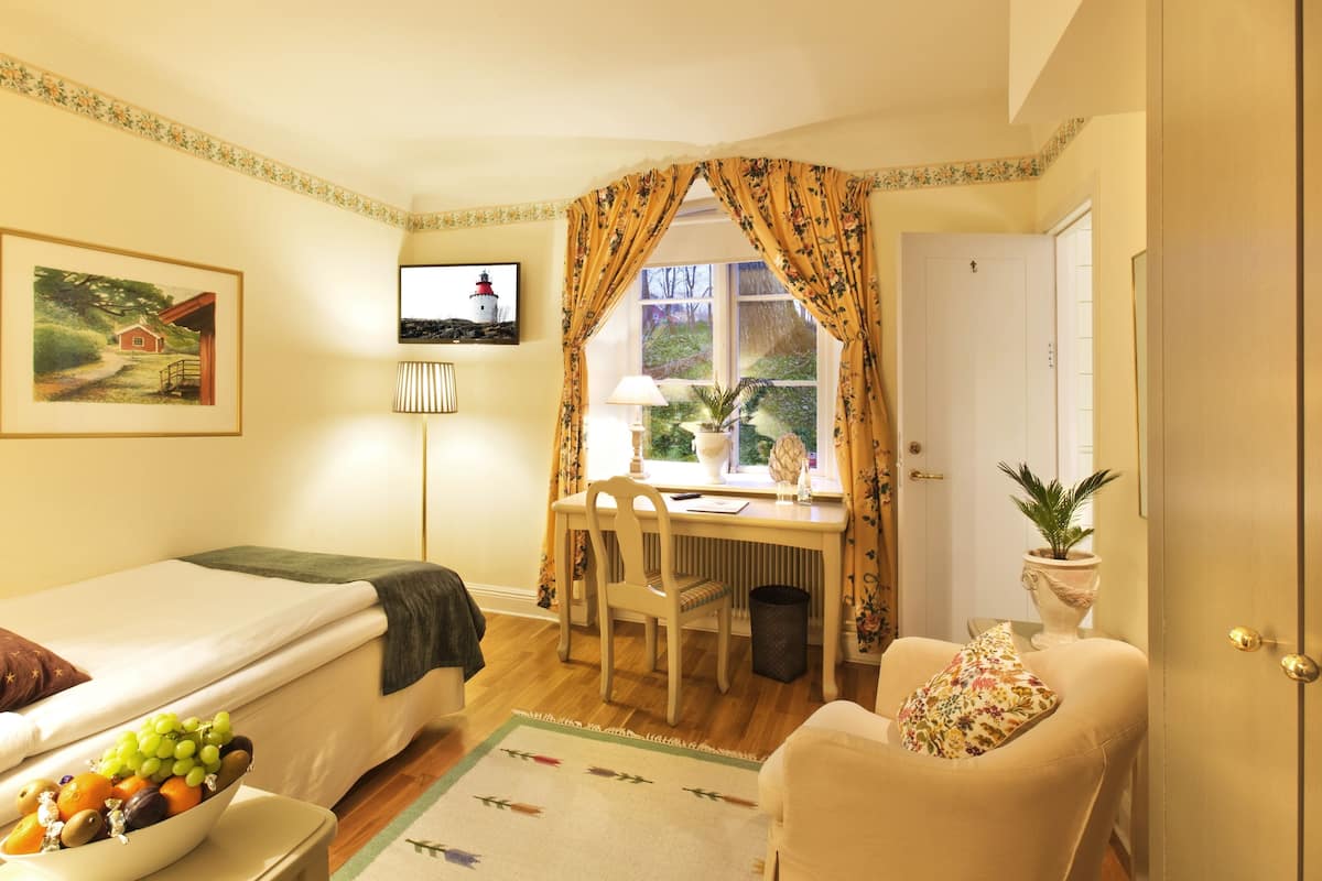 Una habitación acogedora con una cama, un escritorio con una silla, una mesita con un frutero y una ventana con cortinas.