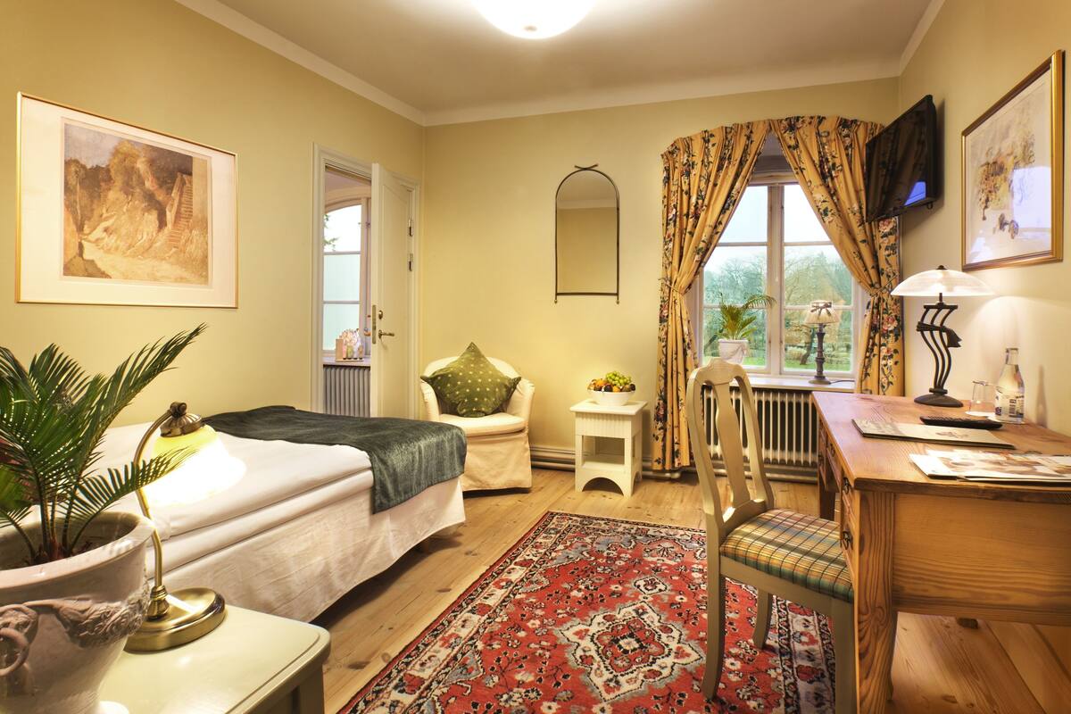 Un dormitorio con cama, mesita de noche, lámpara, silla y vistas al exterior.