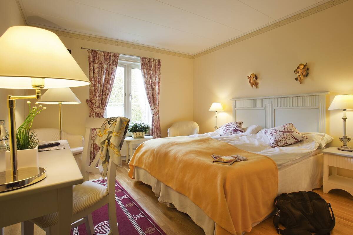 Habitación de hotel con cama, lámparas de noche, escritorio con lámpara, una silla, ventana con cortinas y una mesita con planta.