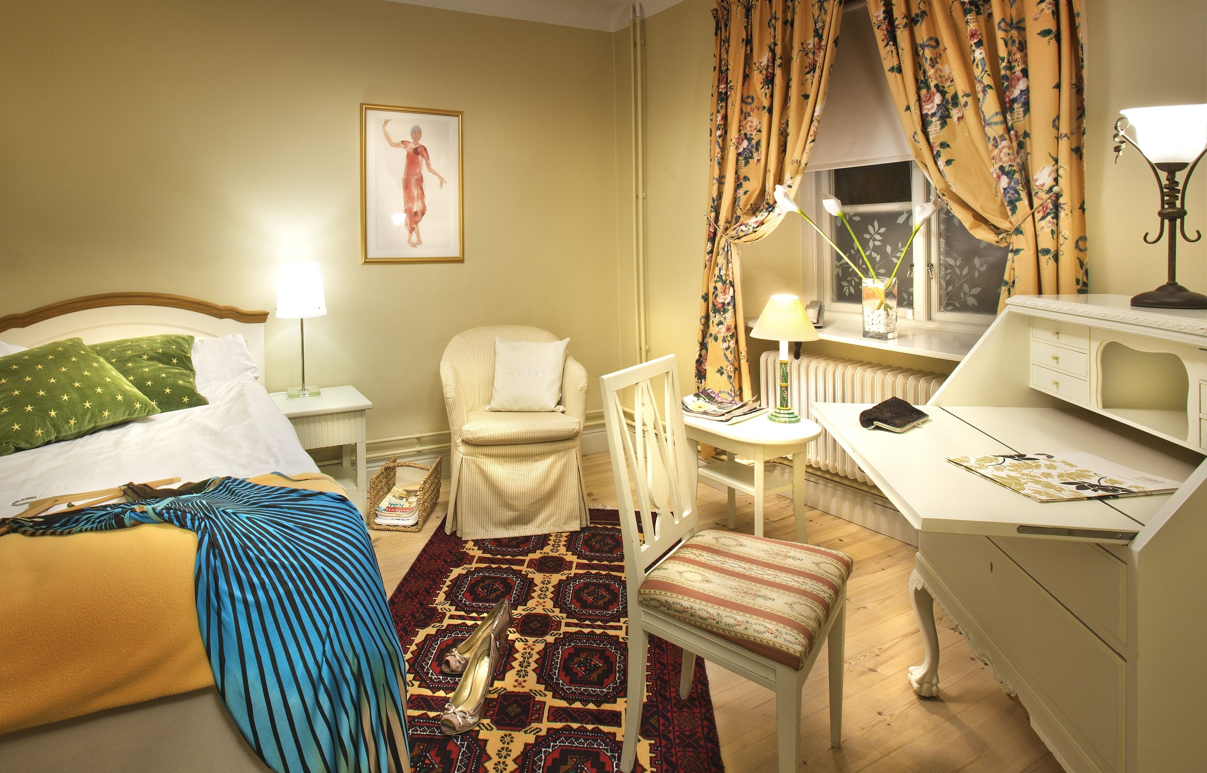 A hotel room with a bed, a desk, a chair, a small table, a lamp, and a window with curtains.