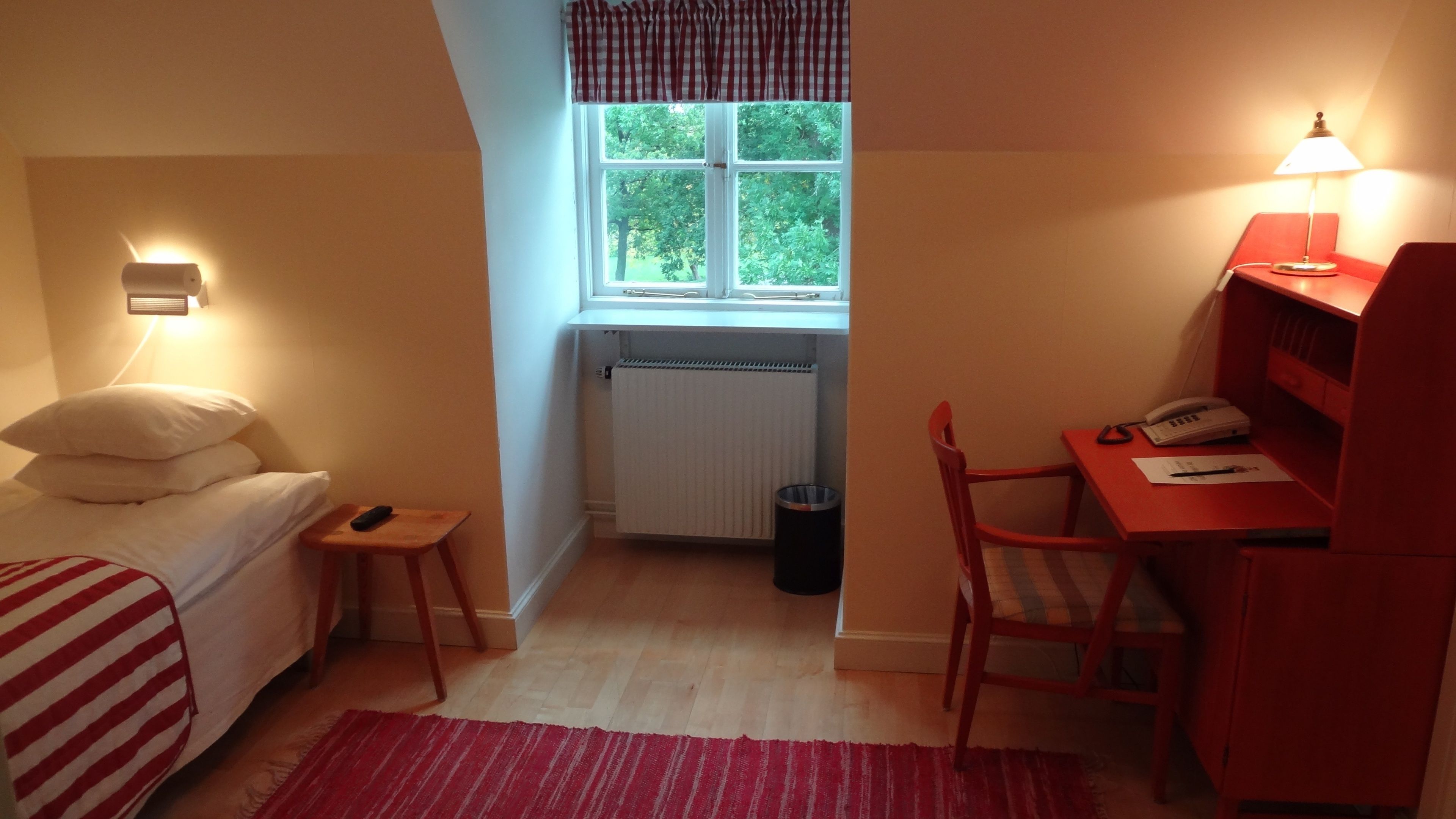 A small, well-lit bedroom with a bed, a desk, and a chair. There is a window with a view of trees outside, a radiator, and a trash bin.