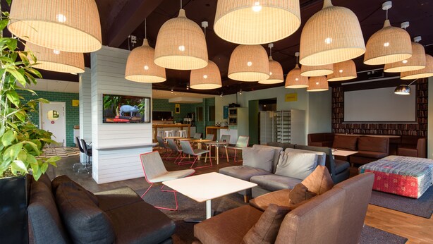 Et moderne loungeområde med flere siddepladser, herunder sofaer og lænestole, belyst af store vævede pendellamper.