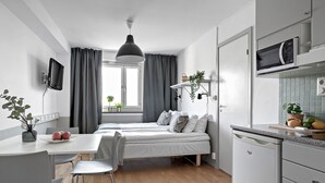 Et moderne, kompakt soveværelse med seng, sengeborde og et skrivebord med stol. Der er et vindue med gardiner, en hængende plante og en mikrobølgeovn i et køkkenområde.