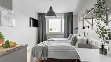 A modern bedroom with a bed, bedside table, and a desk with a chair. There is a window with curtains, a hanging plant, and a mounted TV.