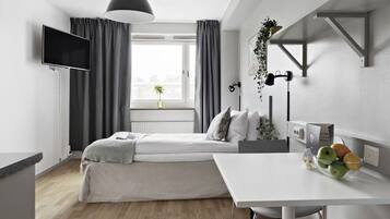 Een moderne slaapkamer met een bed, nachtkastje en een bureau met stoel. Er is een raam met gordijnen, een flatscreen-tv aan de muur en een potplant op het bureau.