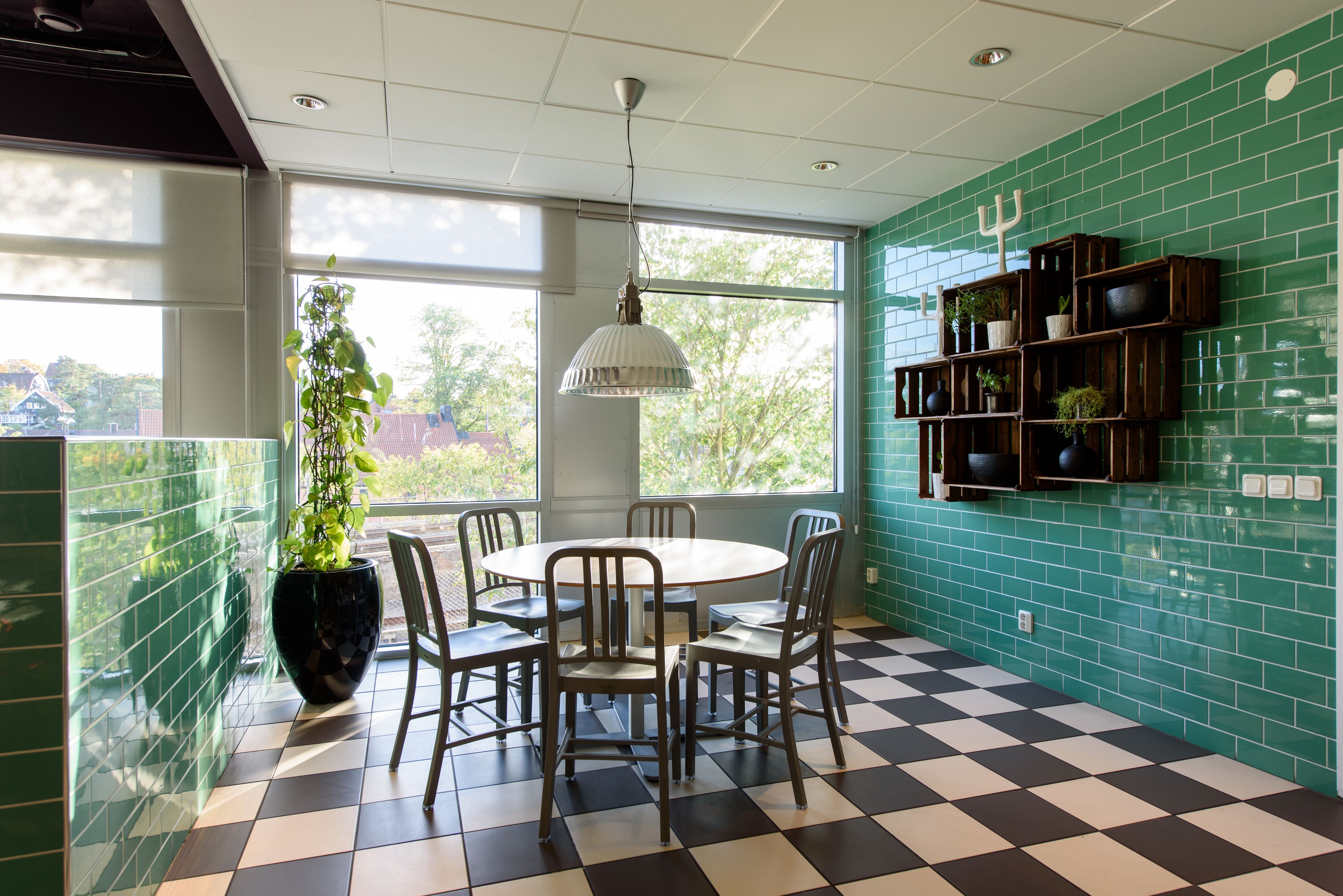 Uma sala de jantar com piso quadriculado, uma mesa redonda e cadeiras. Há um vaso de plantas, um lustre e uma parede com prateleiras contendo plantas e objetos decorativos.