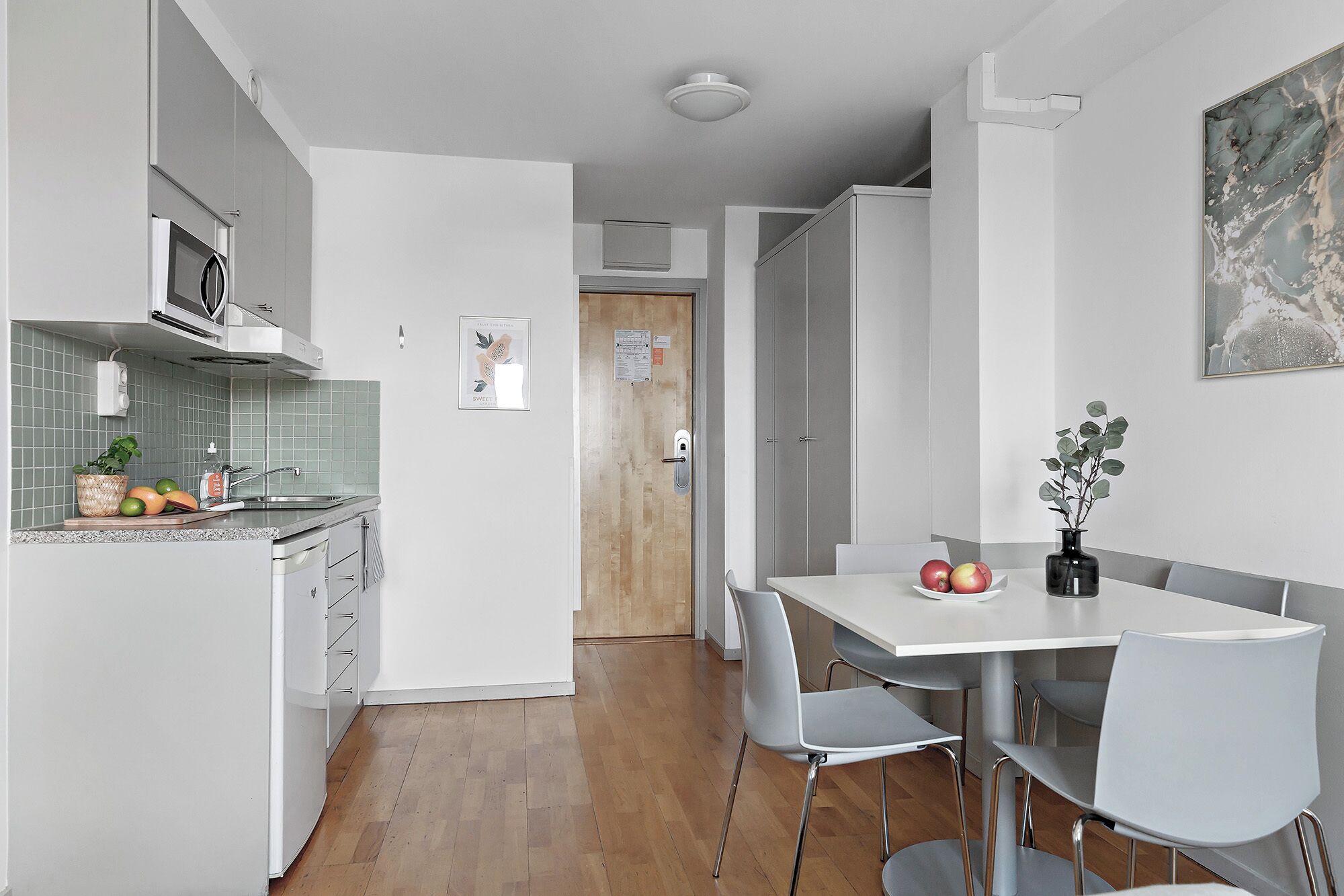 Una cocina moderna con armarios blancos, microondas y salpicadero de azulejos. Hay un comedor con mesa y sillas blancas, y un florero con una planta sobre la mesa.
