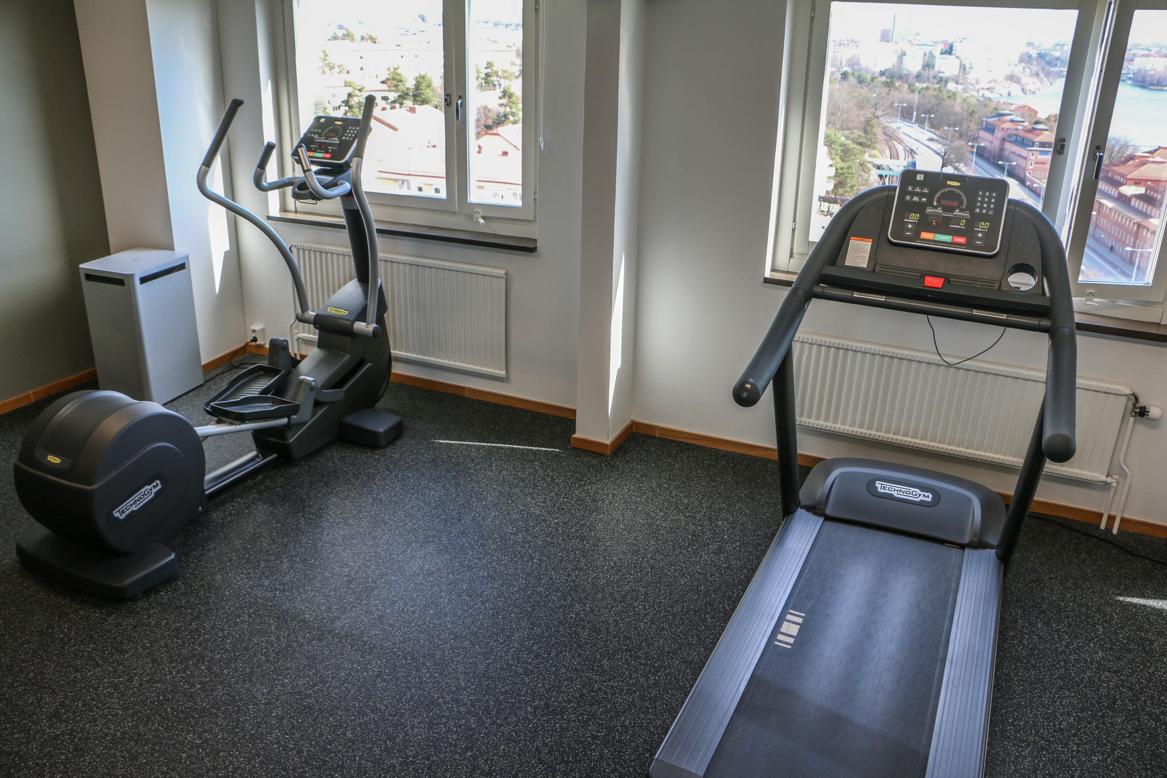 Ein Fitnessraum mit Crosstrainer, Laufband und Fenster mit Stadtblick.