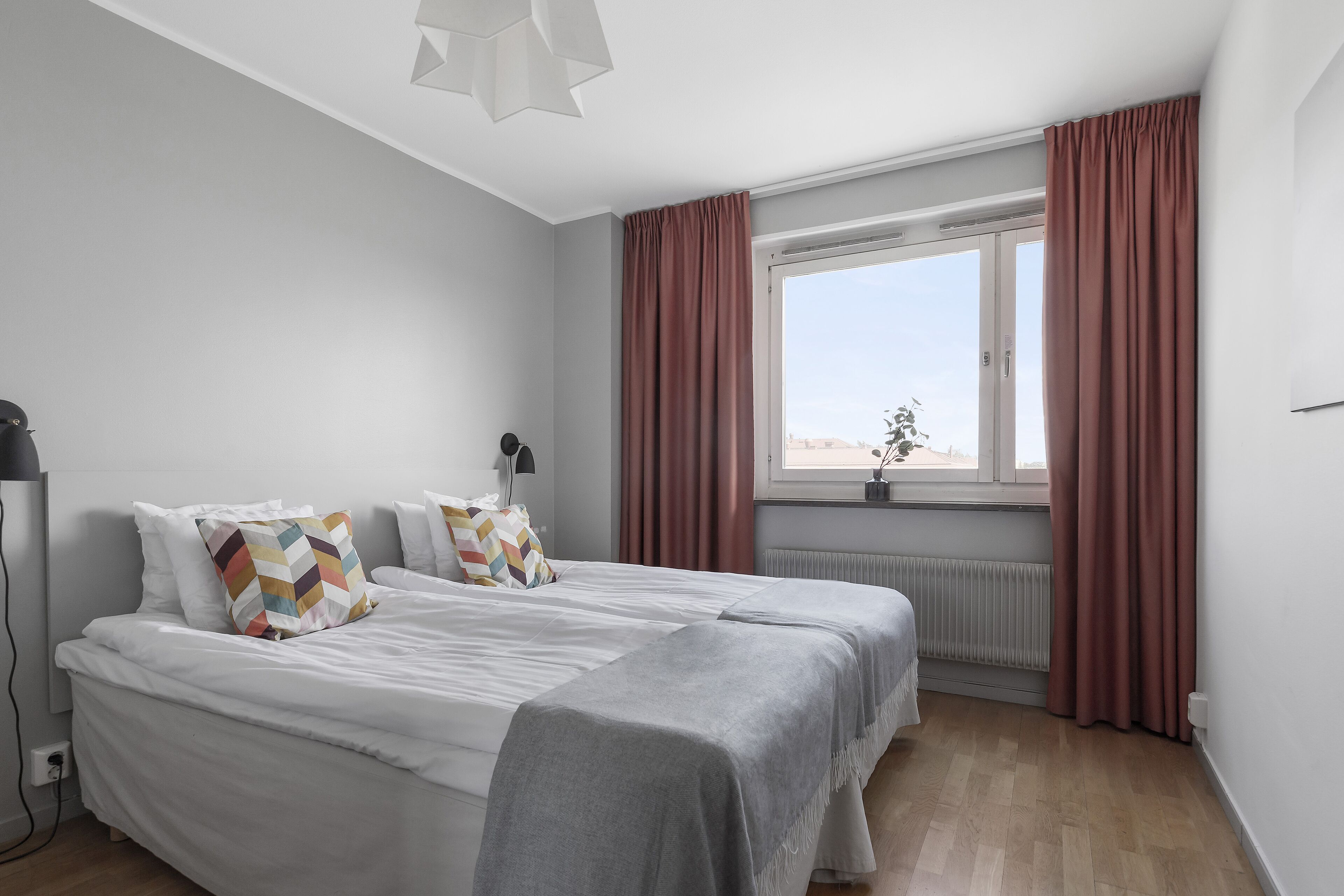 Un dormitorio con cama, ventana con cortinas, radiador y una lámpara fijada a la pared.