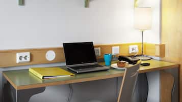 Ein Hotelzimmer mit Schreibtisch aus Holz, einem Laptop, einer blauen Tasse, einem Telefon und einer Lampe.