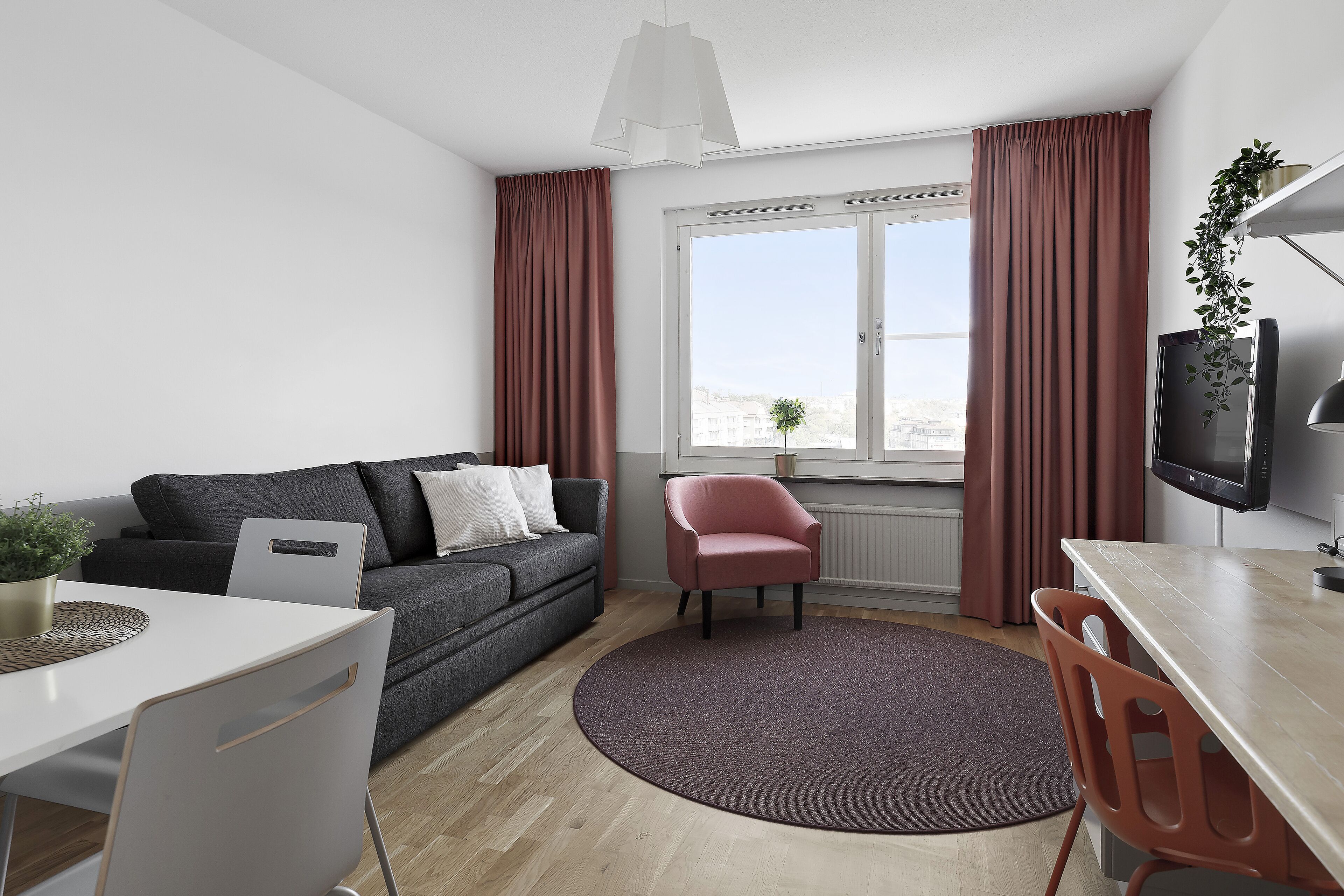 Una sala de estar moderna con un sofá gris, una pequeña silla rosa, una mesa de comedor y un amplio ventanal con cortinas.