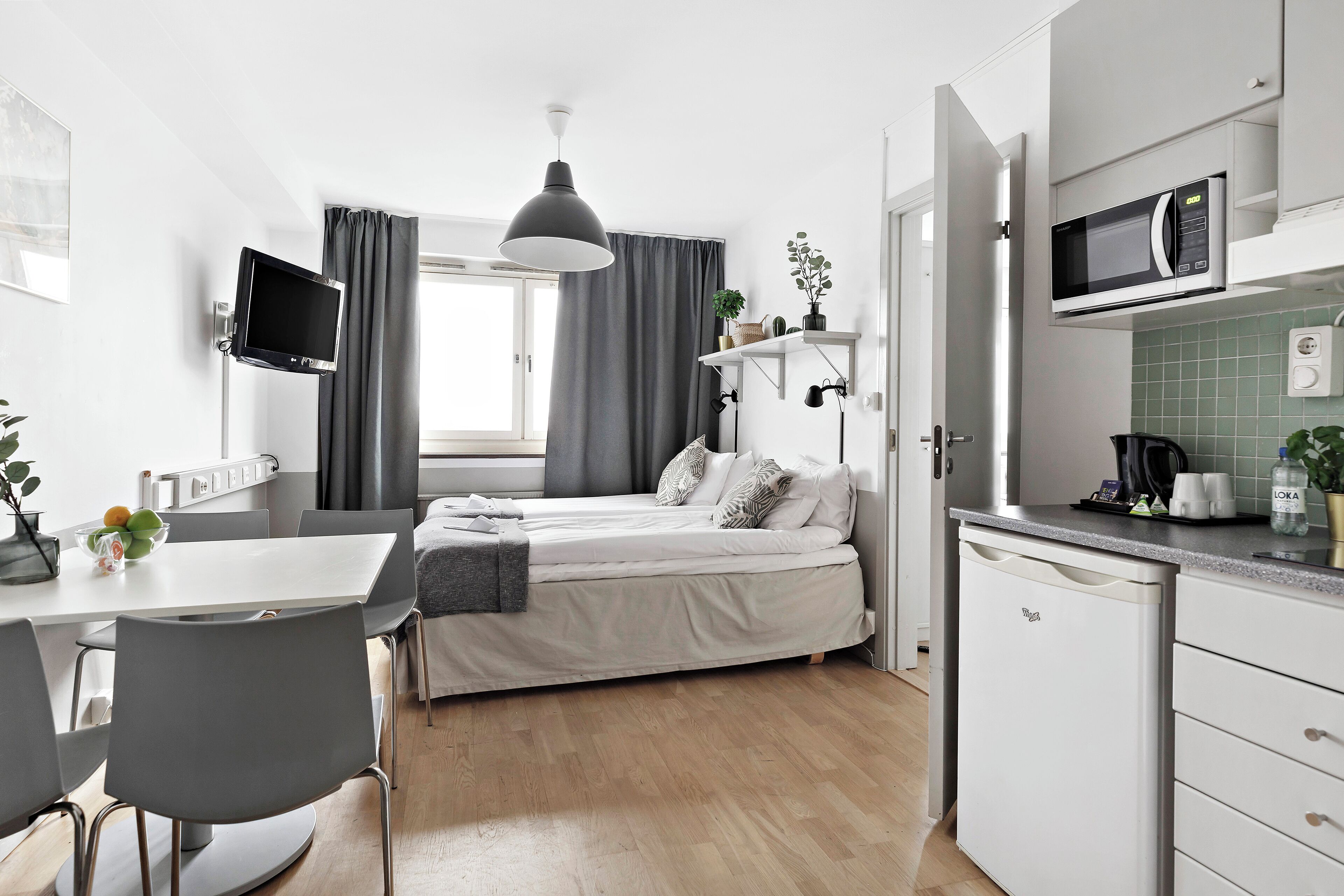 Una habitación de hotel compacta con cama, una mesa de comedor con sillas, un televisor montado en la pared, un microondas y una pequeña cocina.