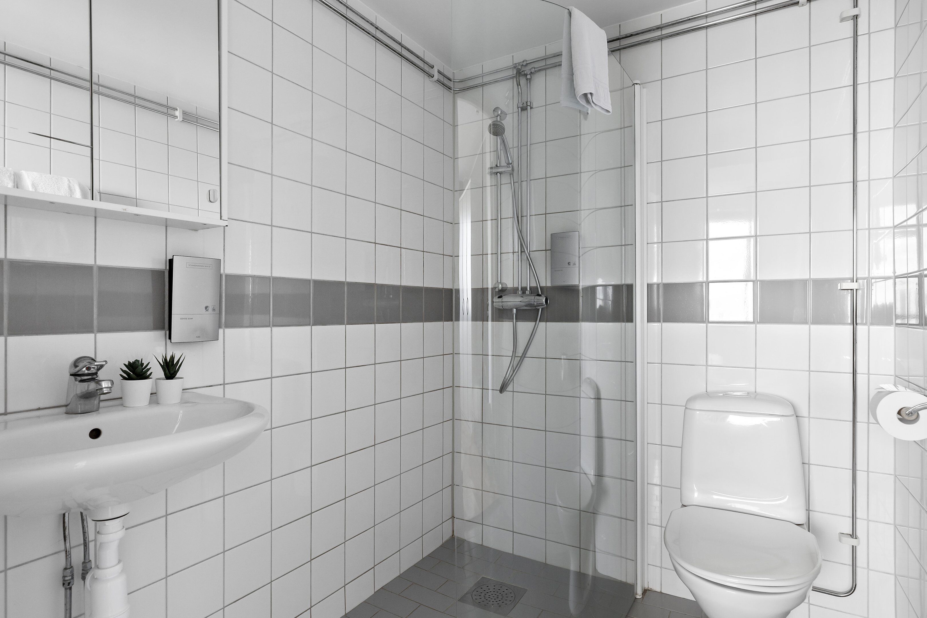 Ein modernes Badezimmer mit weißen Fliesen, einer Duschkabine, einem weißen Waschbecken und einer Toilette.