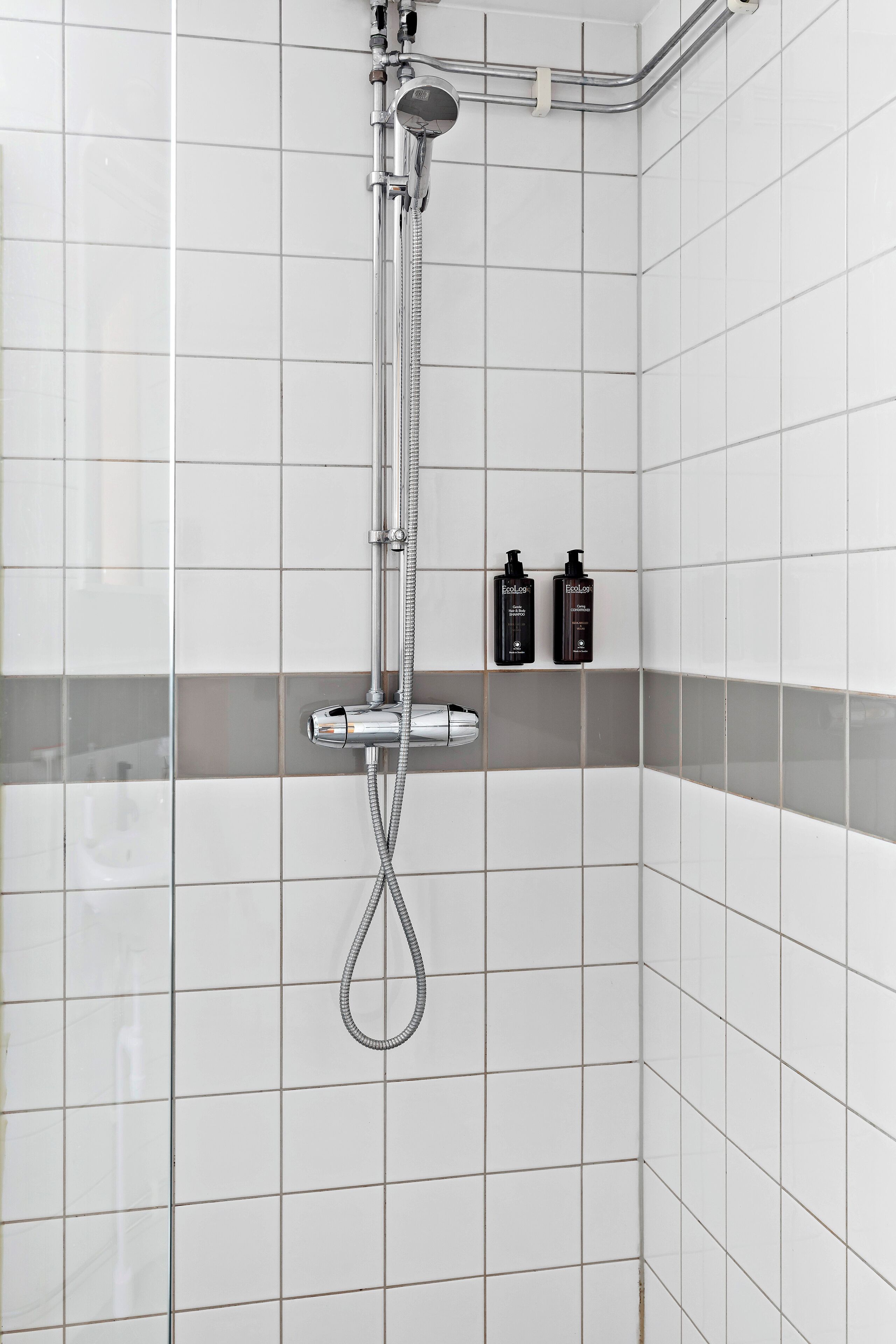 En dusch med ett handhållet duschmunstycke och en väggmonterad blandare. Två flaskor schampo står på en hylla under duschen.