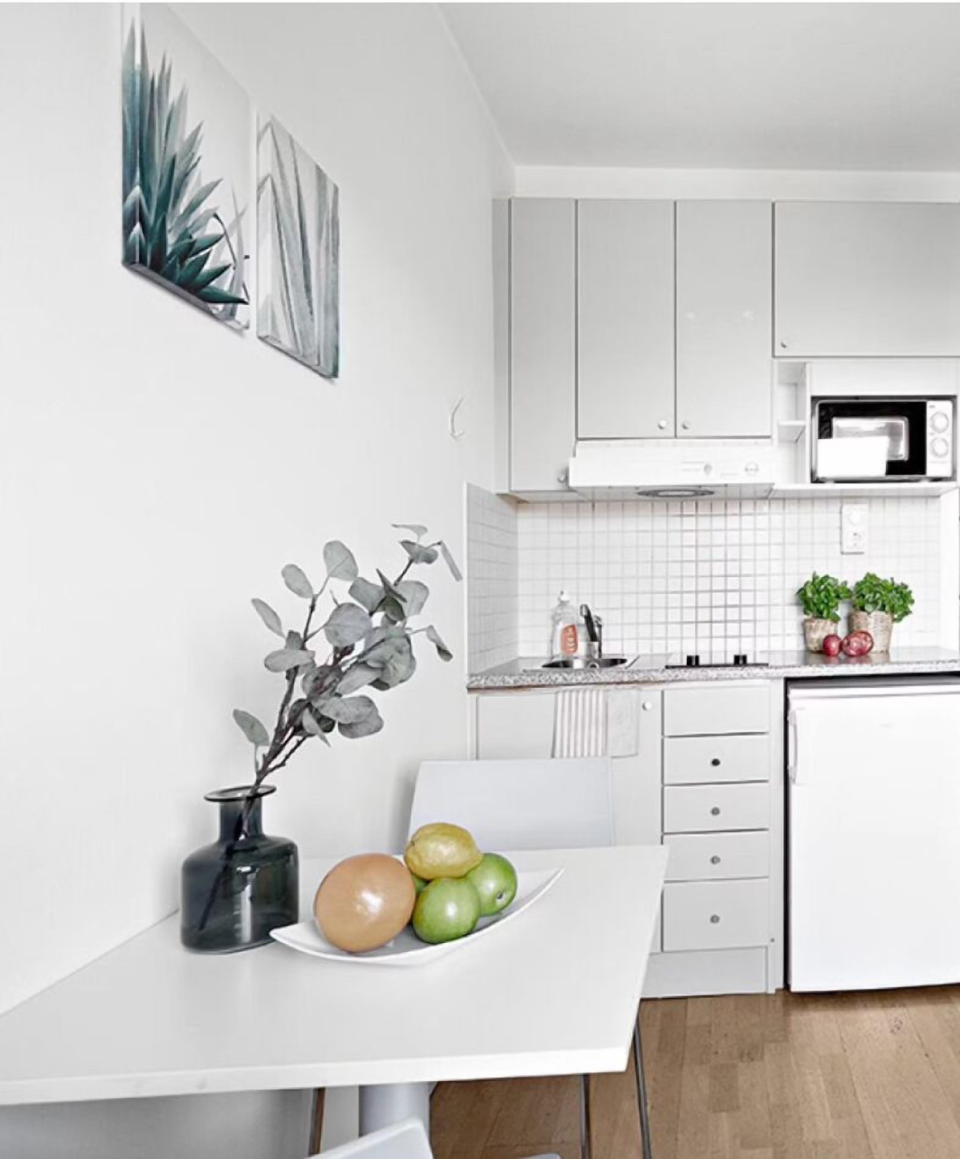 A modern kitchen with white cabinets, a microwave, and a white countertop. There is a vase with a plant on the counter and a plate with apples and a pear.