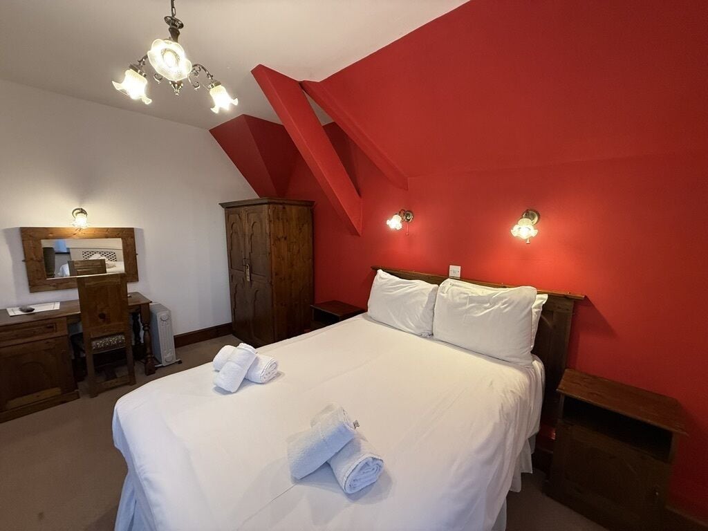 Un dormitorio con cama, ropa de cama blanca, un armario de madera, un escritorio con silla y una pared de acento roja.