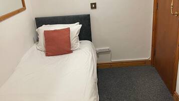 Una cama individual con ropa de cama blanca y roja, un cabecero oscuro, una mesita de noche pequeña y piso cubierto de alfombra.